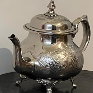 Peut inclure: Une théière argentée avec un couvercle et une anse incurvée. La théière a un bec verseur et repose sur trois petits pieds. Le corps de la théière est orné d'un motif floral en relief.