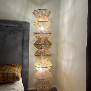 Może przedstawiać: Wysoka, beżowa lampa podłogowa o konstrukcji przypominającej klepsydrę. Lampa wykonana jest z tkanych włókien naturalnych i posiada wiele źródeł światła. Lampa stoi obok łóżka z szarym wezgłowiem i poduszką w kratę. Wysokość ok. 150 cm.