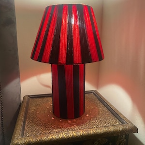 Può includere: Una lampada da tavolo decorativa con paralume e base a righe rosse e nere. La lampada è posizionata su un tavolo di legno ornato e dettagliato con accenti dorati. Il design della lampada presenta strisce verticali.