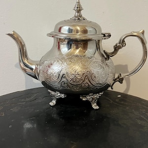 May include: A silver teapot with ornate floral engravings and a decorative lid finial. The teapot has a curved spout and a handle with a decorative design. The teapot sits on three small feet.