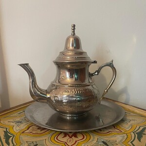 Peut inclure: Une théière argentée avec un bec verseur et une poignée incurvés, posée sur un plateau rond assorti. La théière présente des motifs gravés complexes et un couvercle décoratif. L'ensemble à thé est posé sur une surface colorée et à motifs.