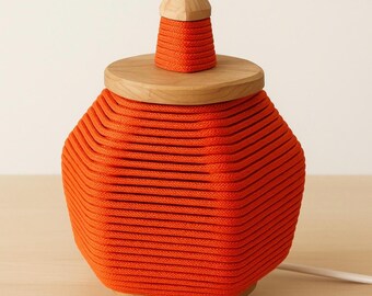 May include: An orange decorative lamp with a wooden lid and a small wooden finial. The lamp's body is made of tightly wound orange rope, and it sits on three small wooden legs. The lamp is plugged into a white electrical cord.