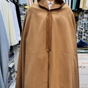 May include: A tan hooded cloak with a zipper closure, displayed on a mannequin. The cloak has a simple design and is made of a soft, flowing fabric. The background shows other clothing items.