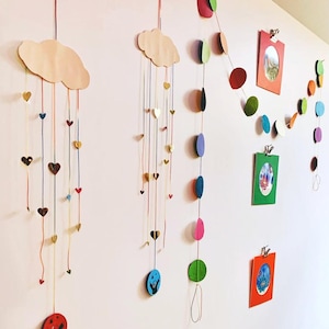 May include: Wall decor featuring wooden cloud shapes with hanging heart and circle ornaments in various colors. Additional hanging garlands of colorful circles and framed photos add a whimsical touch to the wall.