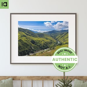 May include: A framed photograph of a lush green valley with a river winding through it. The sky is blue with white clouds. The image is framed in a dark gray frame and has a green sticker that says "REAL IMAGE AUTHENTIC NOT A.I."