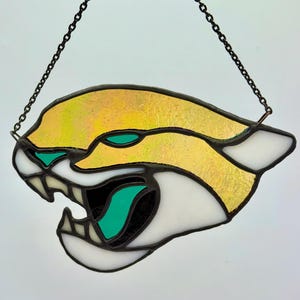 May include: Stained glass cougar head with a fierce expression. The artwork features a yellow and gold body, white accents, and teal eyes and tongue. A black chain is attached for hanging.