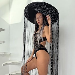 Op de afbeelding: Een zwart bodysuit met kanten details en uitsparingen. Het model draagt zwarte hoge hakken en een grote zwarte hoed met lange franjes. De achtergrond is een witte trap en muur.