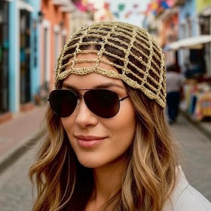 May include: A woman wearing a gold-colored, crocheted head covering and dark sunglasses. The head covering has a net-like design. The woman is outdoors, with a colorful street in the background.