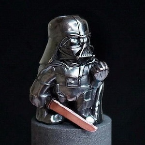 Puede incluir: Figura metálica de Darth Vader con una hoja color cobre, montada sobre una base cilíndrica gris. La figura está detallada con el casco y la armadura del personaje. La palabra "Bimetal" está en la esquina superior derecha sobre un fondo negro.