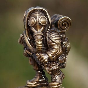Op de afbeelding: Een messing beeldje van een persoon in een gasmasker en een capuchon, met een rugzak. De figuur hurkt op een rots, met details zoals de lenzen van het masker, de opgerolde bovenkant van de rugzak en het woord "Brass".