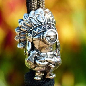 May include: A silver-colored charm depicting a Minion character wearing a Native American headdress and holding a small axe. The charm is attached to a black braided cord. The word "Melchior" is in the upper right corner.