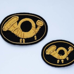 May include: Two embroidered patches featuring a gold-colored horn design on a black oval background. The horn is detailed with intricate stitching, and the patches are set against a white backdrop.