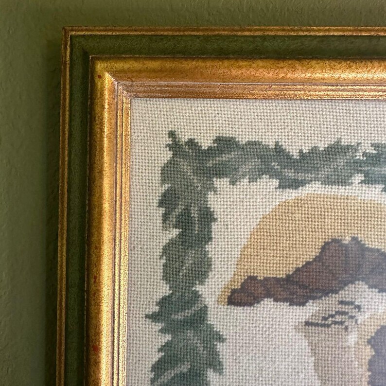 May include: Close-up of a framed needlepoint artwork. The frame has a gold inner edge and a green outer edge. The artwork features a beige background with a green leaf border and a partial image of a figure with brown and tan details.