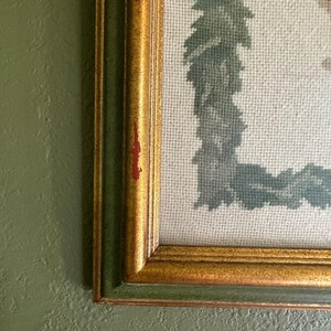 May include: Close-up of a framed needlepoint artwork. The frame has a gold finish with a green inner border. The needlepoint features a light blue and green design on a cream-coloured background. The artwork is mounted on a green wall.