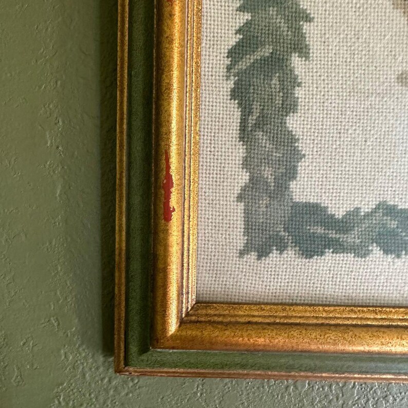 May include: Close-up of a framed needlepoint artwork. The frame has a gold finish with a green inner border. The needlepoint features a light blue and green design on a cream-coloured background. The artwork is mounted on a green wall.