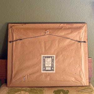 May include: The reverse of a framed picture, featuring a brown paper backing and a black wire for hanging. A small, rectangular label with text and decorative elements is attached to the lower centre of the frame. The frame rests on a patterned surface.