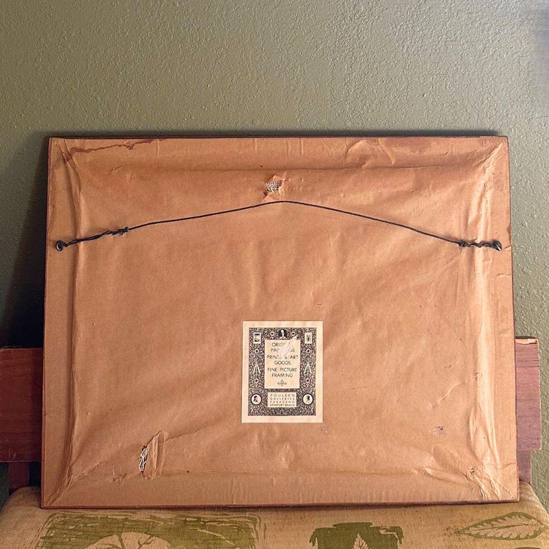 May include: The reverse of a framed picture, featuring a brown paper backing and a black wire for hanging. A small, rectangular label with text and decorative elements is attached to the lower centre of the frame. The frame rests on a patterned surface.