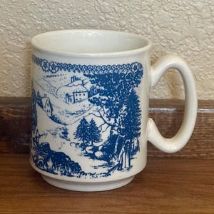 May include: White ceramic mug with a blue landscape design. The design features a winter scene with trees, houses, and figures. The mug has a rounded base and a curved handle.