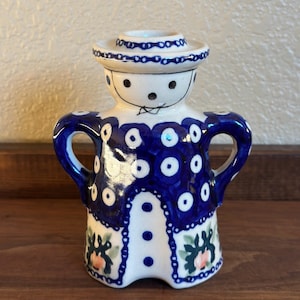 May include: A ceramic figurine in the shape of a person, featuring a white base with blue and white polka dots and floral accents. The figurine has a hat and handles, with a painted face. The color scheme is primarily blue and white.