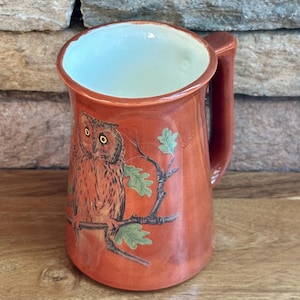 May include: A ceramic mug with a glossy burnt orange finish featuring a hand-painted brown owl perched on a dark tree branch with green oak leaves. The interior of the mug is a smooth off-white color, and it includes a sturdy, matching orange handle.