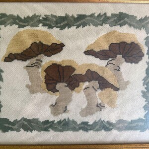 May include: Framed needlepoint art depicting three mushrooms with tan and brown caps and stems, set against a white background. The artwork is framed with a gold-coloured frame and a border of green leaves.