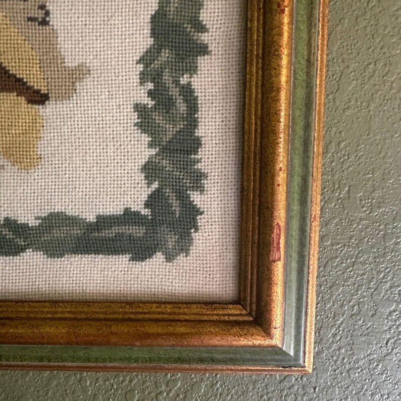 May include: Needlepoint artwork in a gold-toned frame with a green and gold border. The artwork features a light-coloured background with a green wreath design. Part of a yellow and brown figure is visible.