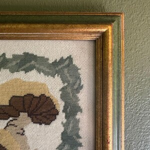 May include: Close-up of a framed needlepoint artwork. The frame is gold with a green outer edge. The needlepoint features a floral design with brown, tan, and green colours. The artwork is displayed against a green wall.