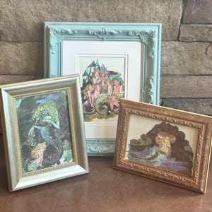 May include: Three framed art prints featuring mermaid illustrations. The frames are in various colors, including light blue, gold, and silver. The artwork depicts mermaids in a fantasy setting with vibrant colors and detailed imagery.