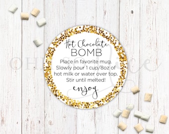 May include: A round, white sticker with a gold glitter background and black text that reads "Hot Chocolate BOMB". The text below reads "Place in favourite mug. Slowly pour 1 cup/8oz of hot milk or water over top. Stir until melted! enjoy"