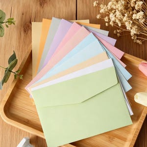 Könnte beinhalten: Eine Sammlung pastellfarbener Umschläge, die auf einem Holztablett aufgefächert sind. Die Umschläge sind in Gelb-, Orange-, Grün-, Rosa- und Blautönen gehalten. Ein Strauß Trockenblumen und eine kleine dekorative Säule befinden sich im Hintergrund.