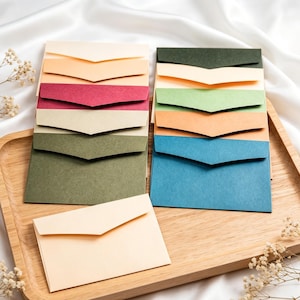 Puede incluir: Una colección de sobres pequeños y coloridos en varios tonos, incluyendo crema, rojo, verde y azul, dispuestos sobre una bandeja de madera. Los sobres están cuidadosamente apilados, mostrando su diseño plegado. El fondo presenta una tela blanca y sedosa.