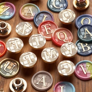 May include: A collection of wax seal stamps with gold-colored letters and plaid patterns. The seals are in various colors, including pink, blue, and red. The letters of the alphabet are featured on the stamps.