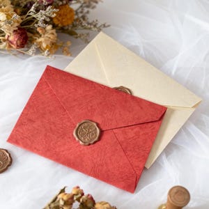 May include: Two envelopes, one red and one cream, sealed with wax stamps. The red envelope has a textured surface and a gold-colored wax seal. Dried flowers are in the background, suggesting a vintage or handcrafted theme.