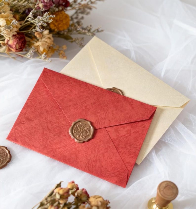 May include: Two envelopes, one red and one cream, sealed with wax stamps. The red envelope has a textured surface and a gold-colored wax seal. Dried flowers are in the background, suggesting a vintage or handcrafted theme.