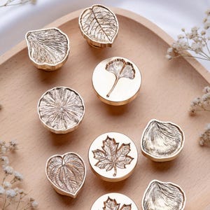 Può includere: Una collezione di timbri per ceralacca color oro con vari disegni botanici, tra cui foglie e un ginkgo. I timbri sono disposti su un vassoio di legno chiaro, mostrando i loro dettagli intricati e la finitura metallica. Ideali per artigianato e cancelleria.