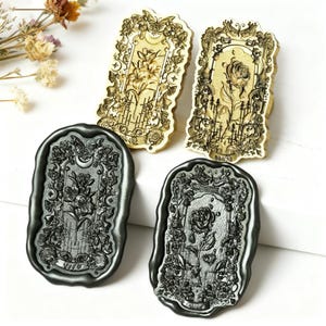 May include: Four decorative wax seal stamps. Two are gold-toned with floral and butterfly designs, and two are dark gray with floral and rose motifs. The stamps are rectangular with ornate borders, ideal for crafting and stationery.