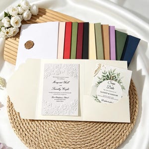 Könnte beinhalten: Ein cremefarbenes, dreifach gefaltetes Hochzeitseinladungs-Set mit einer weißen, geprägten Blumenkarte und einer Save-the-Date-Karte mit grünen Blattillustrationen. Das Set enthält eine Auswahl an farbigen Umschlägen in Weiß, Rot, Grün, Schwarz und Blau.