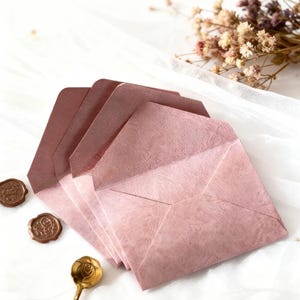 May include: A stack of pink textured envelopes with a matching spoon and two wax seals. The envelopes are arranged on a white cloth, with dried flowers in the background. The envelopes are a dusty rose color.