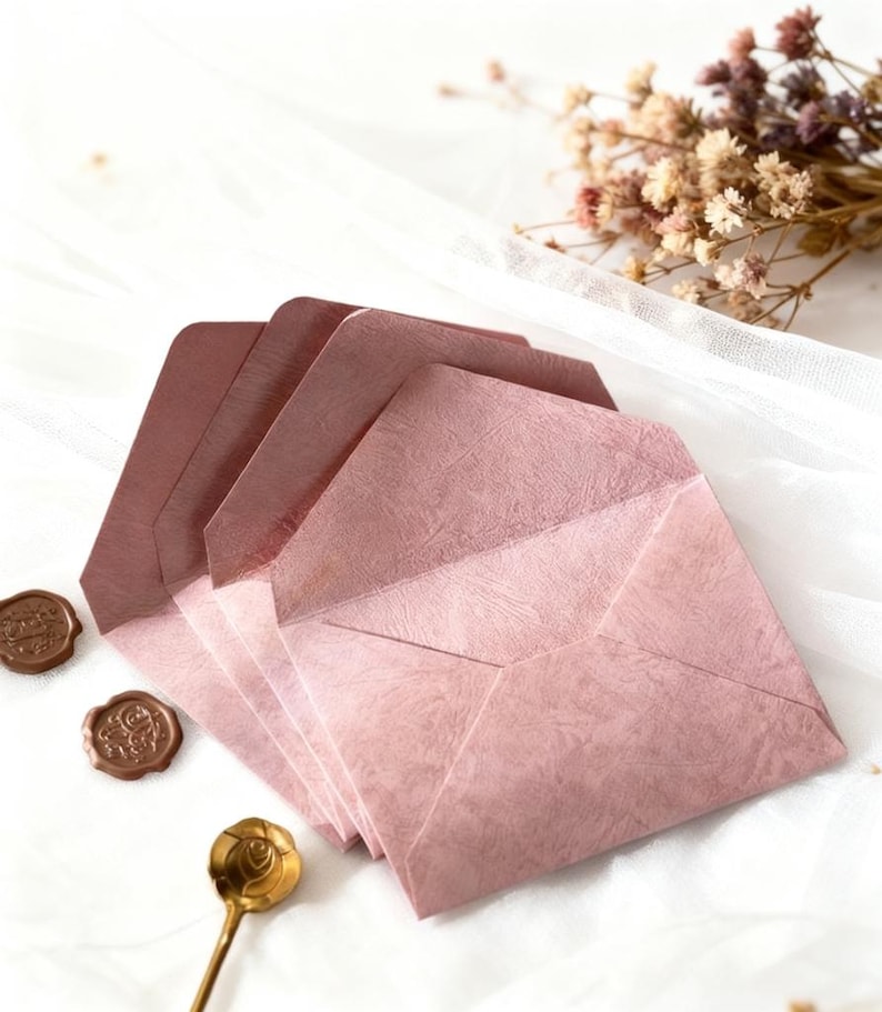 May include: A stack of pink textured envelopes with a matching spoon and two wax seals. The envelopes are arranged on a white cloth, with dried flowers in the background. The envelopes are a dusty rose color.