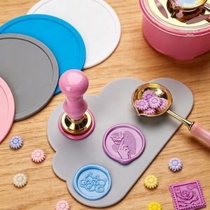 Puede incluir: Kit para hacer sellos de cera con un sello rosa, una cuchara con cuentas de cera moradas y varios sellos de cera de colores. Los sellos presentan diseños florales y de mano sosteniendo una rosa. También se ven posavasos redondos.