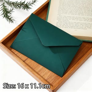 May include: A dark green envelope, measuring 16 x 11.1 cm, sits in a wooden tray. The envelope is closed and has a simple design. The tray is made of light-coloured wood. A sprig of greenery and a book are in the background.