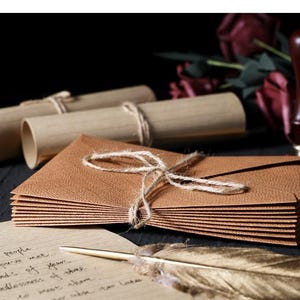May include: A stack of brown textured envelopes tied with twine, alongside rolled scrolls and a feather quill. A wooden stamp and burgundy flowers are in the background. A handwritten letter is visible in the foreground.