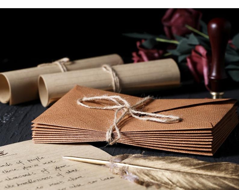 May include: A stack of brown textured envelopes tied with twine, alongside rolled scrolls and a feather quill. A wooden stamp and burgundy flowers are in the background. A handwritten letter is visible in the foreground.