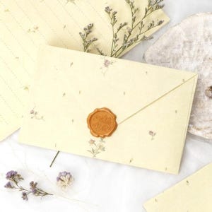 May include: Cream-coloured stationery set with floral accents. The envelope is sealed with a gold wax stamp. The set includes an envelope, lined paper, and additional stationery, all adorned with delicate flower illustrations.