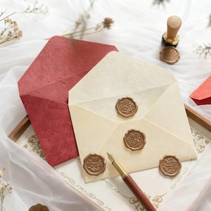 May include: Two envelopes, one red and one cream, are displayed with decorative wax seals. A pen with a brown barrel and gold accents rests near the envelopes. A wax seal stamp is also visible, suggesting a stationery or correspondence theme.