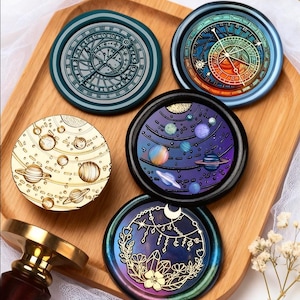 Puede incluir: Una colección de sellos de cera decorativos con diseños celestiales y astronómicos. Los sellos presentan planetas, constelaciones y patrones intrincados de relojería en varios colores. También se ven una bandeja de madera y un sello.