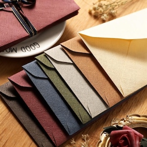May include: A collection of textured envelopes in various colors, including brown, beige, blue, and green. Some envelopes are stacked, while others are fanned out. A plate with the words "GOOD MORNING" is visible.