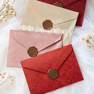 May include: An assortment of colorful envelopes with wax seals. The envelopes are in shades of red, pink, and cream, with a textured finish. Each envelope is sealed with a detailed, circular wax seal. The envelopes are arranged on a white, textured surface.