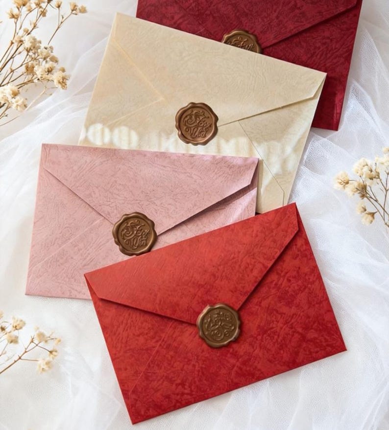 May include: An assortment of colorful envelopes with wax seals. The envelopes are in shades of red, pink, and cream, with a textured finish. Each envelope is sealed with a detailed, circular wax seal. The envelopes are arranged on a white, textured surface.