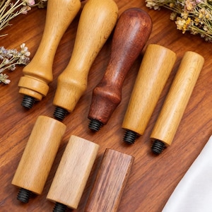 May include: A collection of wooden stamp handles in various shapes and shades of brown. The handles have a threaded base for attaching stamps. The wood grains and textures are visible, with some handles featuring a darker, reddish-brown hue.